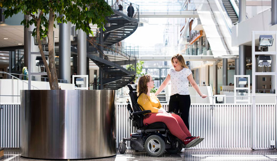 Ung kvinne i rullestol sammen med assistent i atrium i skole