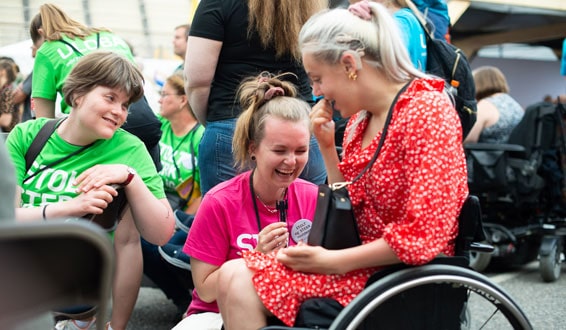 Kvinne med sommerkjole i rullestol ler sammen med assistent blant mange mennesker
