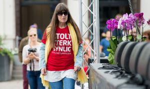 Berit Vegheim går mot kamera langs scenen på Independent Living-festivalen 2016. Ho ser bestemt ut og har på seg raud T-skjorte med "Stolt, sterk, synlig" på brystet, gult skjerf og blå olabukse. Langs scenekanten er det lilla og rosa blomar. 