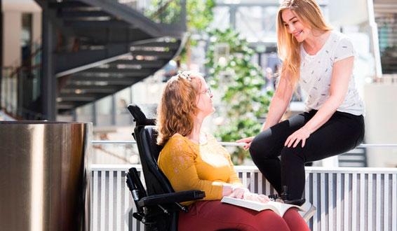 Ung kvinne i rullestol med assistent i høyskolebygg