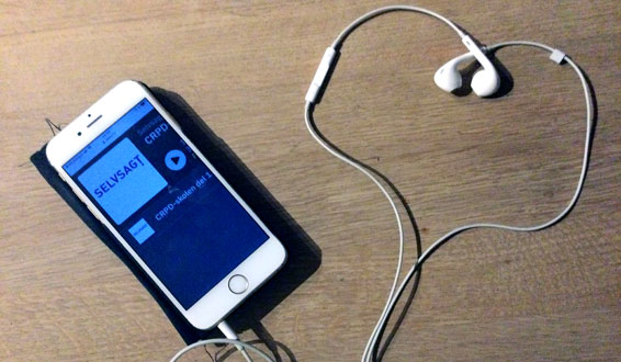 Mobiltelefon som viser Selvsagtpodden i displayet, ørepropper med ledninger som former et hjerte.