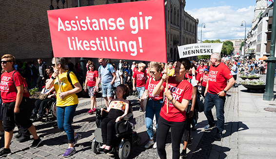 Et illustrasjonsfoto fra Stolthetsparaden 2016. Mennesker i røde T-skjorter med "Stolt sterk synlig" på brystet marsjerer nedover Karl Johan. Noen av dem bærer en stor fane der det står "Assistanse gir likestilling".