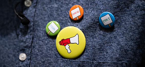 Et blått skjortebryst med fire buttons på, En grønn meden plakat og ordet "sterk", en tilsvarende oransje med ordet "synlig" og en blå med ordet "stolt". Den siste buttonen er større en de andre og er gul med bilde av en rød og hvit ropert.