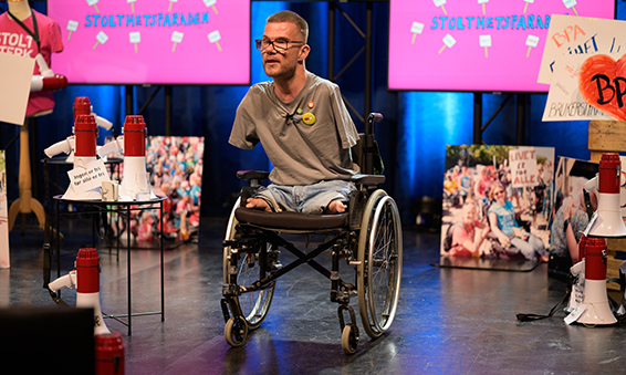 Bilde av Sebastian Tjørstad på settet til Stolthetsparaden 2021 på Verket scene i Moss. Sebastian sitter i en manuell rullestol, han mangler armer og ben, og har på seg olabukse og grå T-skjorte med Independent Living festivalbuttons på brystet. Han har briller, kortklippet hår og litt skjegg. I bakgrunnen er kollasjer av bilder fra tidligere Stolthetsparader, skjermer med den rosa Stolthetsparaden-illustrasjonen på, megafoner med små kort med paroler og slagord festet til, og større plakater.