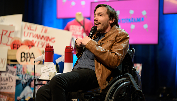 Bilde av Johannes Ramstad Finnanger i manuell rullestol, sort bukse, grå T-skjorte og brun skinnjakke som gjør stand up på digital Stolthetsparade 2021. I bakgrunnen er den rosa Stolthetsparadeillustrasjonen, plakater, megafoner og bannere.