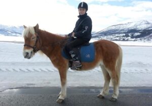 Arbeidsleder Regine Elvevold sittende på en hest i hvitt snølandskap. Regine har blondt hår under ridehjelmen, svart jakke, bukse og rutete sokker. Hun sitter på en lysebrun hest. 