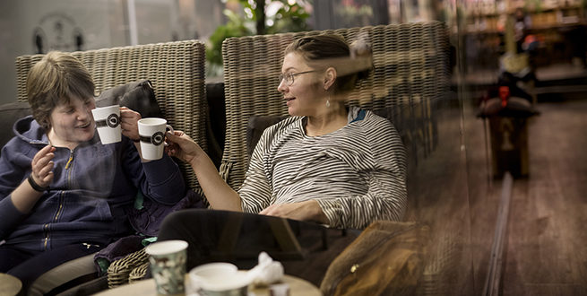 Live Spiten og assistenten sitter på kafé og skåler med hver sin kaffekopp. Bildet tas gjennom et vindu, og man får derfor en speileffekt på glasset. Live har på seg blå hettejakke, har kortklippet lysebrunt hår og smiler mot assistenten. Assistenten har sort- og hvitstripet genser, håret i en topp på bakhodet, lange øredobber med blanke stener i, og briller.