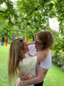 Bilde av Guro Helene Sørdalen sammen med kjæreste Simen Bondevik fra partiet Sentrum. De to står i en hage omgitt av grønt gress og planter og holder rundt hverandre. Man ser en lenke av pride-flagg i bakgrunnen.