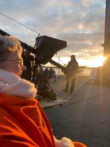 Sonja på jubileumskonsert med KORK i Bø i Vesterålen. Hun har en sprek, oransje anorakk på seg, hvitt skjerf, og kveldssola i ansiktet. I bakgrunnen vises konsertdeltagere.