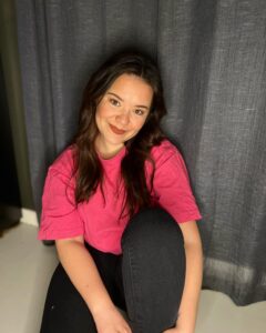 Guro Helene Sørdalen i knallrosa T-skjorte og mørke jeans. Hun sitter på gulvet foran det som ser ut som en gardin, og smiler.