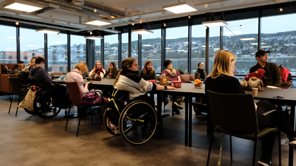 Møteromet Skansgård på Independent Living-huset, har glasveggar slik at ein får utsikt mot Drammen. Søndag 4. desember samla dei 15 deltakarane seg rundt det grå langbordet der inne for å lære om ulike aktivistiske uttrykksmåtar. Biletet viser bordet med deltakarane rundt, og glasveggane od utsikta i bakgrunnen.