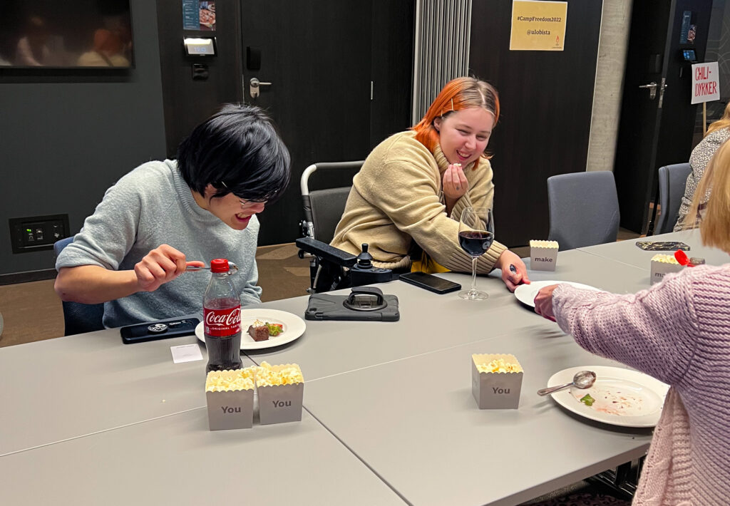 Hanna Betten og Thu Ha Le ler medan dei et kake og snakkar med andre deltakarar. Dei feirer 3. desember. Hanna har off-white genser og oransje hår med litt ettervekst, Thu har lysblå genser og svart bobklipp. På bordet framfor henne ligg paden med taleverktøyet ho brukar.  