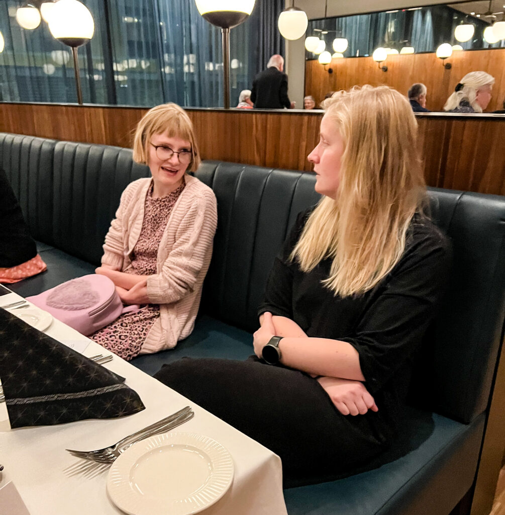 Ingrid Bjørnstad i samtale med Henri Nielsen under middagen 3. desember. Ingrid har lys rosa kjole med leopardmønster, lys rosa cardigan og lys rosa sekk, kort blond bob med pannelugg og briller. Henri sit vendt mot henne i svart kjole og med det blonde litt-over-skulderlange håret laust.