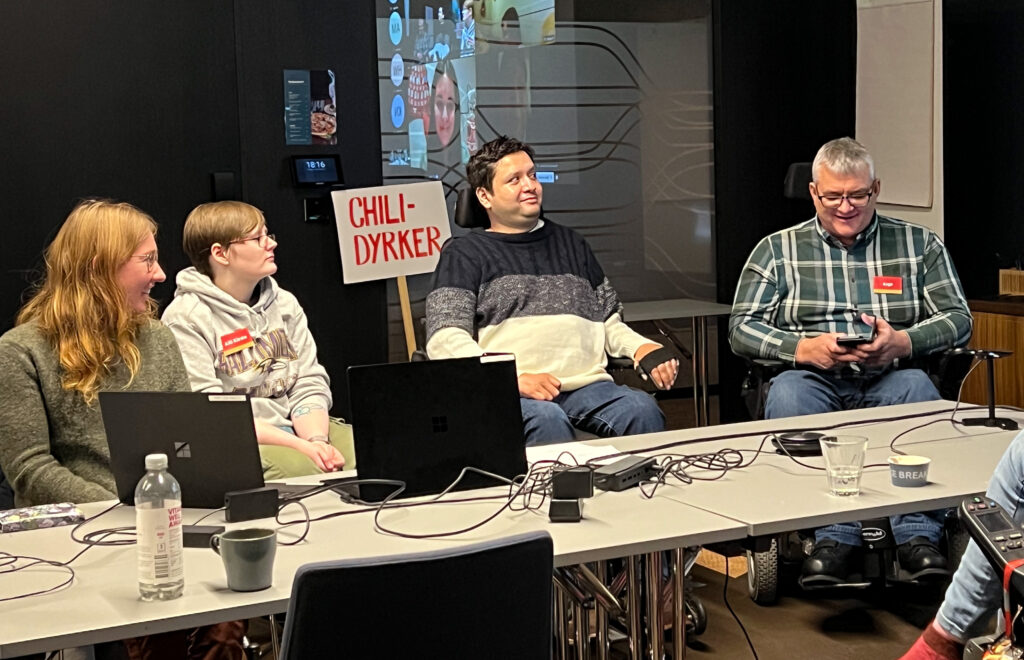 Martine Eliasson, Aili Kirste Arntzen, Jo Benjamin Iversen og Aage Gjesdal i panelsamtale. Arntzen har kort mellomblondt hår, lysgrå hettegenser og briller, Iversen har svart hår, genser i ulike blå-nyansar og sit i elektrisk rullestol. Aage Gjesdal har raud namnelapp, grått hår, briller og rutete skjorte. Han ser ned på talepunkta på mobilen sin. Bak Jo Benjamin står ein parole frå ein tidlegare stolthetsparade, med påskrifta "Chili-dyrker".