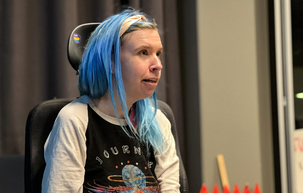 Camp Freedom-deltakar Rina Wesenberg deltek i diskusjonen. Hen har blått, skulderlangt hår, genser med trykk av bandet Journey og lys rosa hårband. Ho sit i elektrisk rullestol. 