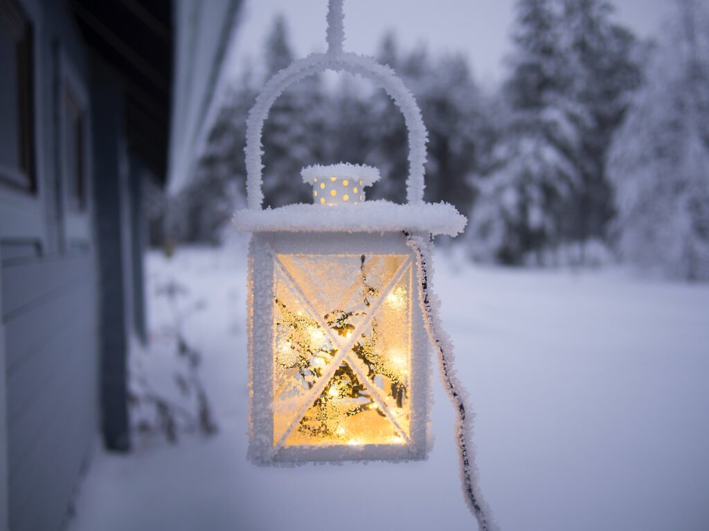 En lykt med varmt lys henger utenfor et snødekt hus. Det er snø på bakken, på trærne og langs den grå husveggen. Lykten er dekket med snø og is, og det varme lyset skinner imot oss.