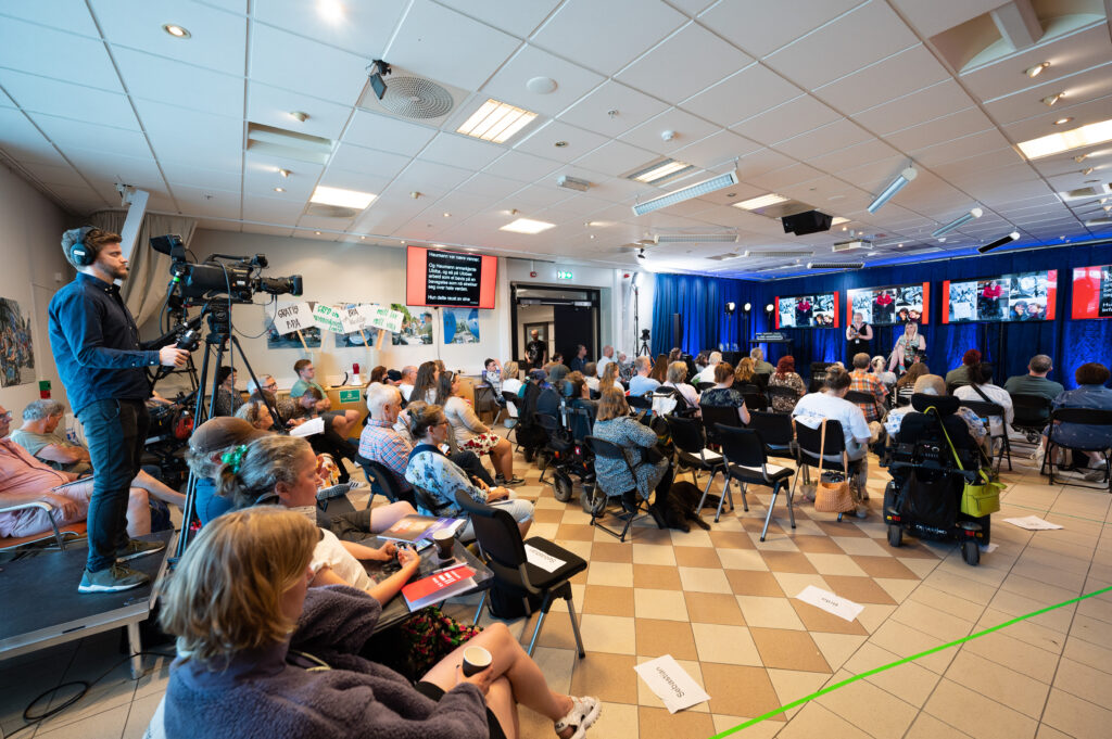 Et overblikk over rommet Roberts, der Lilkestillingsbrølet ble holdt. Vi ser rommet bakfra med scenen, publikum og kameramannen som filmer det hele. På scenen står konferansierene Rina Wesenlund og Mone Celin Skrede. Rina har blondt langt hår og har på seg en lysegrønn kjole med mange tegninger på, som symboliserer funkisaktivisme. Hen sitter i en elektrisk rullestol og har glitrende joggesko. Mone har på seg en svart jumpsuit, har blå øyne og har barbert hodet sitt. Hun har en septum-piercing i nesen og en arm som er mindre enn den andre. Hun har svart øyesminke. På den ene armen har hun en midlertidig tatovering med teksten "Stolt, sterk, synlig!", i tillegg til buttons i rødt, gult og blått med Ulobas slagord på. På scenen ser vi mange store skjermer, samt scenelys og en skjerm med teksttolkning.