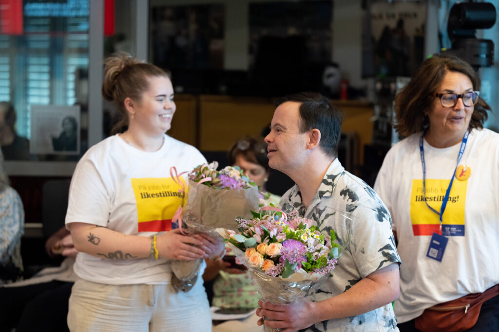 Skuespiller Svein André Hofsø mottar blomster fra en assistent. Han smiler bredt mens han får blomstene, som er rosa, gule, grønne, hvite og sommerlige. Han har mørkebrunt hår og hvit skjorte med trykk. I bakgrunnen ser vi Uloba-ansatt Hilde Lygr med brunt hår og briller.