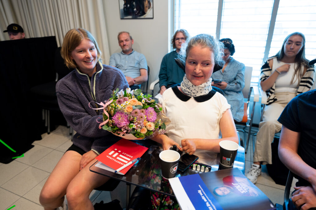 Filmprodusent Mari Storstein med blomstene hun har fått. Hun sitter i en elektrisk rullestol og har blondt hår, skjerf og hvit t-sjorte med svart krage. Foran henne ligger beslutningsstøtte-rapporten "Mitt liv, mitt valg" til Uloba og en blokk med Likestillinsbrølet-logoen. Blomstene er rosa, gule, hvite og sommerlige. Rundt Mari sitter det mange mennesker i publikum.