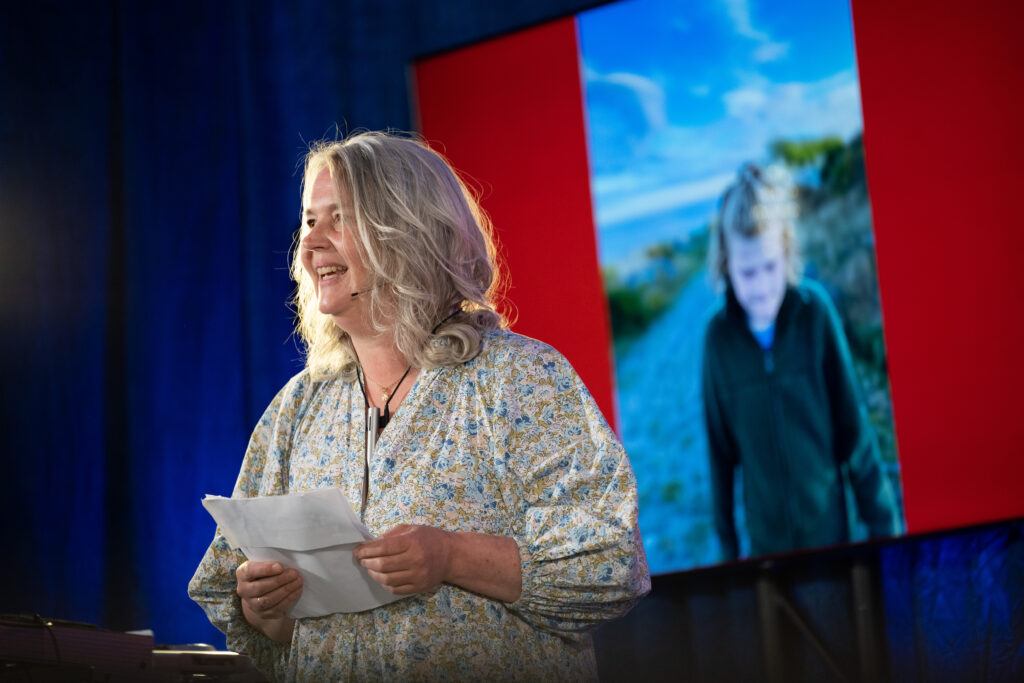 Elisabeth Skredlund står på scenen og forteller om sin datter Alma, som vi ser på skjermen bak henne i en film som moren har laget. Elisabeth har på seg en blomstrete kjole i hvitt, grønt og blått og har blondt hår. Hun leser opp en tekst som hun holder foran seg. Hun har mikrofon og et stort smil om munnen.