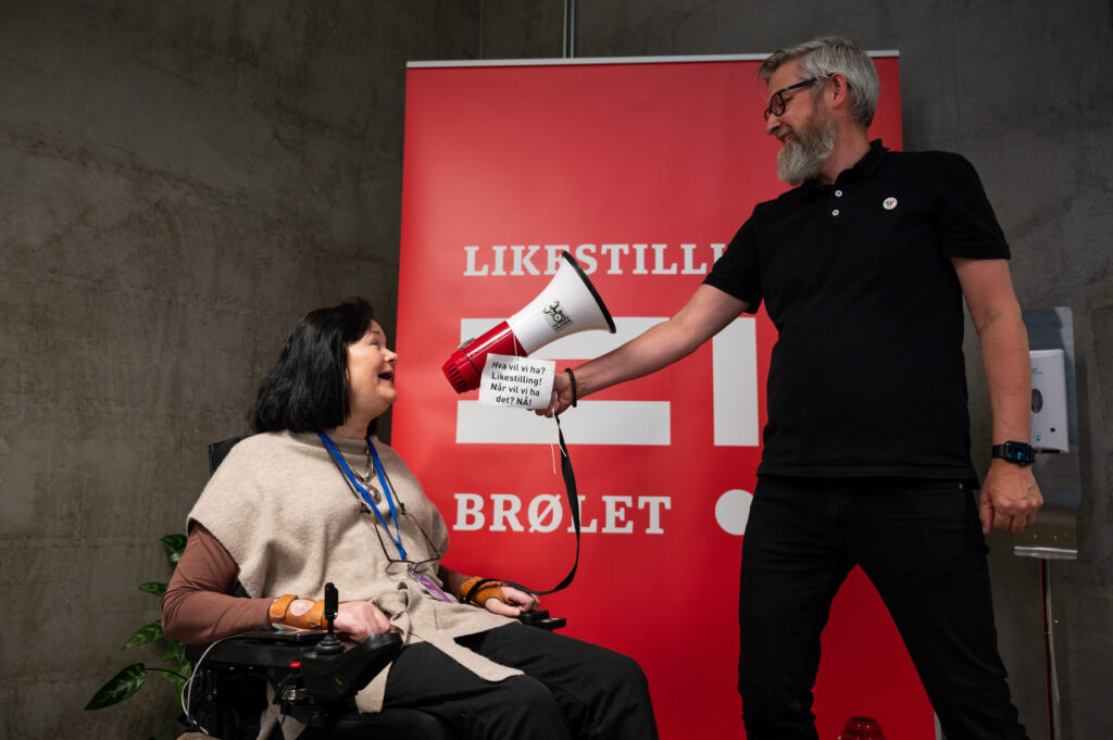 Generalsekretær Vibeke Marøy Melstrøm sammen med Cato Brunvald Ellingsen foran en rød rollup med Likestillingsbrølet-logoen på. Han holder en rød og hvit ropert foran munnen til Vibeke. På den er det festet en lapp der det står "Hva vil vi ha? Likestilling! Når vi vil vi ha det? Nå!". Vibeke har skulderlangt brunt hår og sitter i en elektrisk rullestol mens hun roper inn i roperten. Hun har også et blått Uloba-bånd rundt halsen. Cato har grått hår og skjegg, svarte briller, svarte klær og smiler bredt.