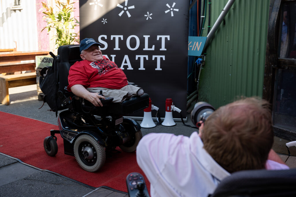 Knut Flåum på rød løper på vei inn i Basar-teltet. Bak ham ser vi en fotovegg med Stolt natt-logoen på, slik som i det forrige bildet av Vibeke. Knut har på seg rød "Stolt, sterk, synlig!"-t-skjorte, beige shorts og mørkeblå caps. Han har grått hår og sitter i en elektrisk rullestol og smiler bredt til Andreas som tar bilde av ham.