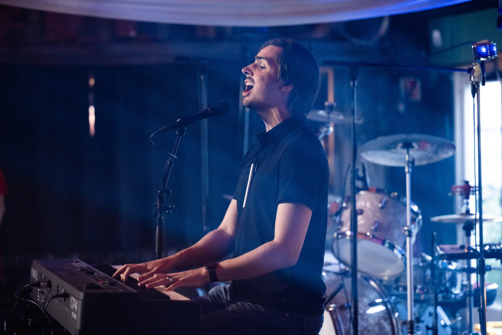 Thomas Tvedt på scenen. Han synger og spiller på et elektrisk piano. Han har mørkebrunt hår, litt skjegg, svart bukse og svart pique-skjorte, samt en svart klokke på armen. Han synger inn i mikrofonen med lukkede øyne og åpen munn. Bak ham ser vi trommer og andre instrumenter.