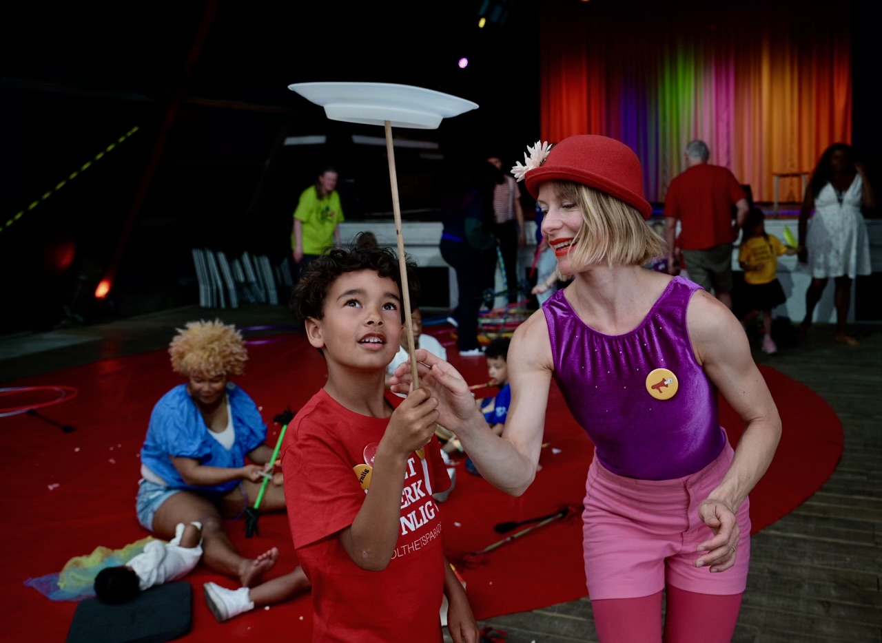 Sirkuskurs med Plutselig Sirkus. En kvinnelig sirkusartist i rosa og lilla antrekk lærer en liten gutt å snurre en tallerken på en pinne. 