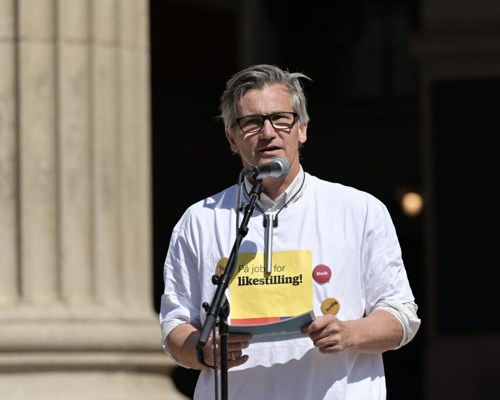 Byråd Bjørn Erik Thon holder appell på Universitetsplassen. Han har kort grått hår og briller, snakker inn i mikrofonen og har på seg en hvit og gul festival-t-skjorte.