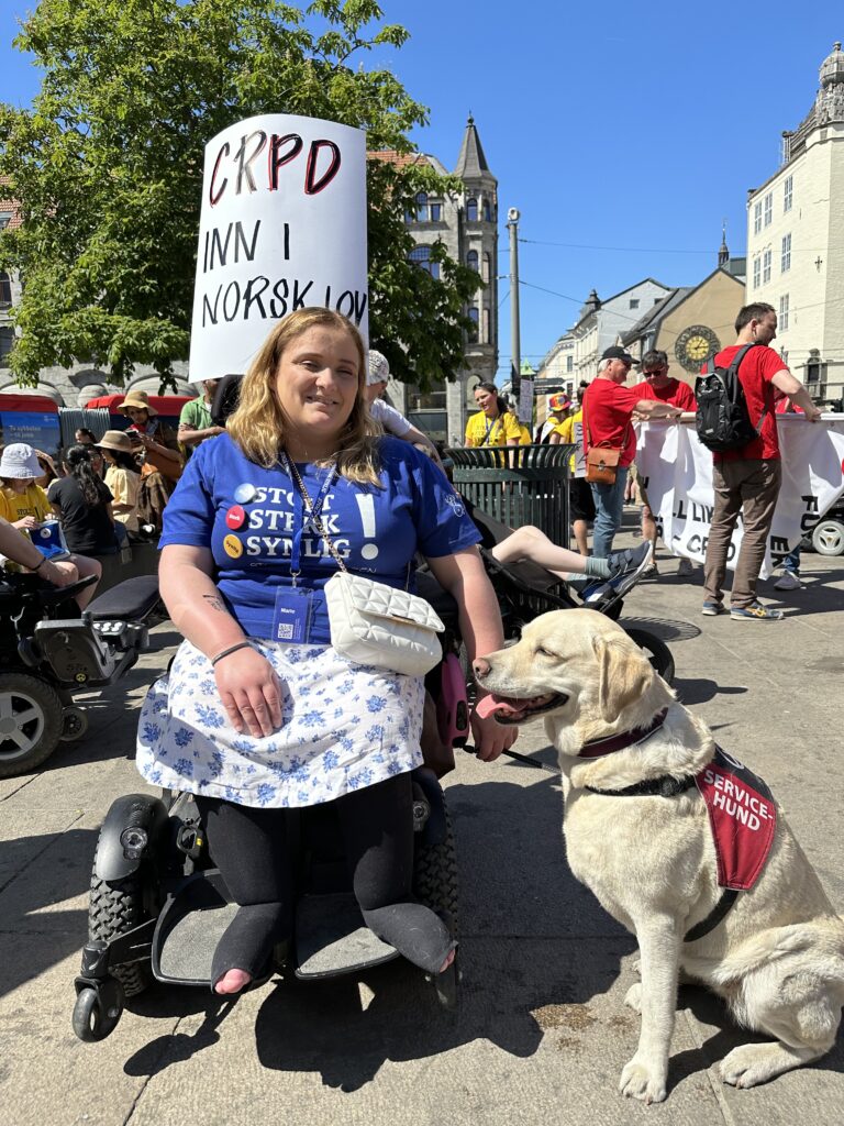 En kvinnelig festivaldeltaker på Universitetsplassen sammen med en hvit labrador som er servicehund. Bak henne ser vi parolen "CRPD inn i norsk lov!". 