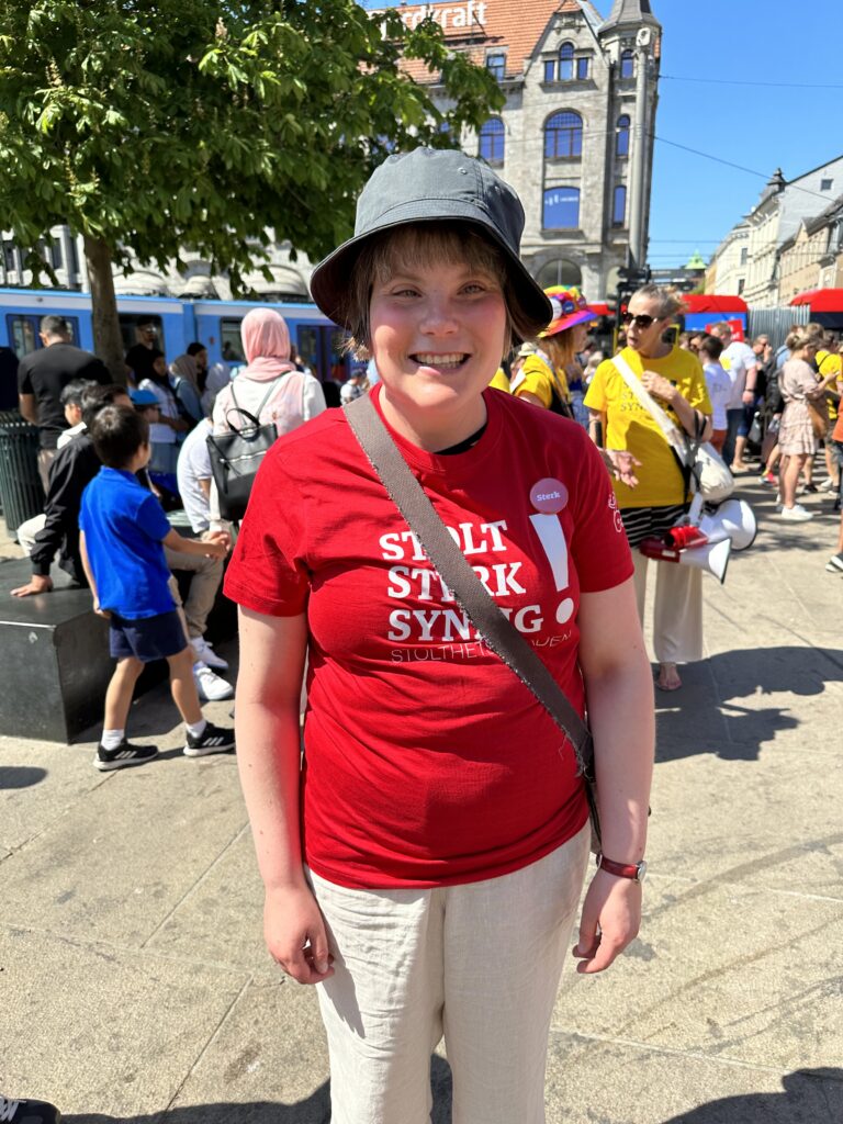 Festivaldeltaker Live Knutsen Spiten på Jernbanetorget etter Stolthetsparaden. Hun er strålende glad og har på seg en rød festival-t-skjorte, en mørk bøttehatt, rød klokke og beige bukse. På t-skjorten er det festet en rød button med teksten "Sterk".
