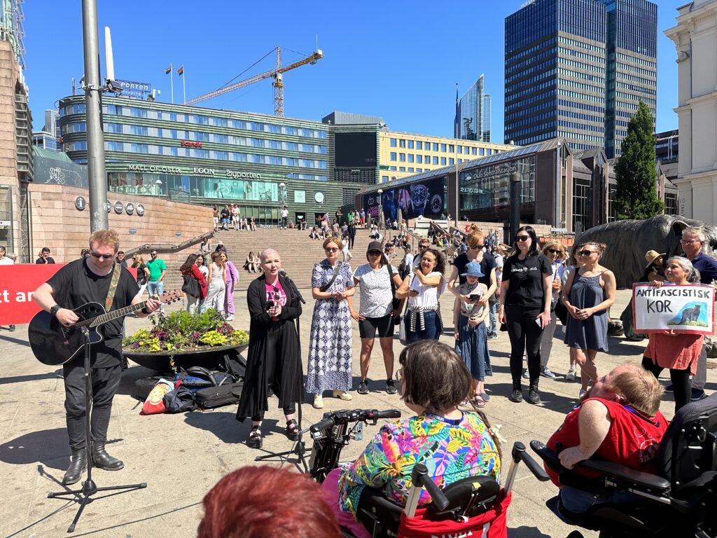 Antifascistisk kor synger for oss på Jernbanetorget. Til venstre står en gitarist i svarte klær, og ved siden av ham står Mone Celin Skrede, programlederen for Likestillingsbrølet, og synger solo. Til høyre for henne står syv andre kordeltakere, alle er jenter. Foran koret står publikum og følger med.