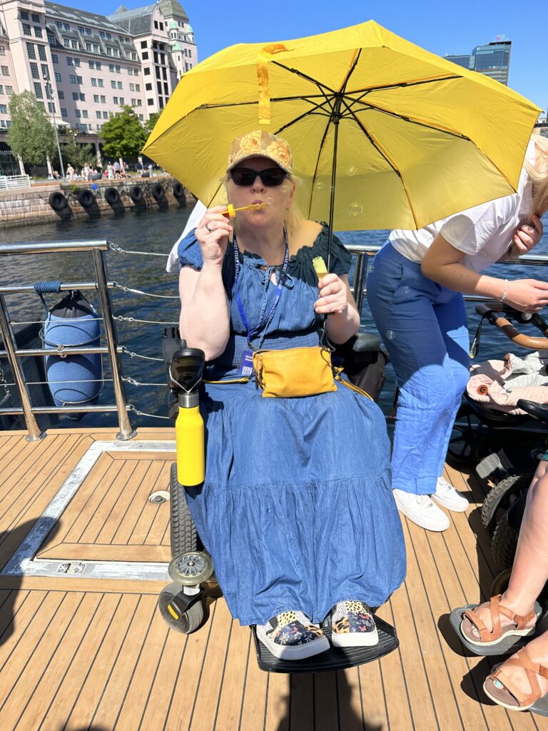 Ulobas May Nina Hansen Auby på båttur med båten Arnøy. Hun sitter i en elektrisk rullestol iført en blå denim-kjole og holder en gul paraply over hodet. Vesken matcher paraplyen. Hun blåser såpebobler og har på seg caps og solbriller. I bakgrunnen ser vi blått hav og havnen på Salt.