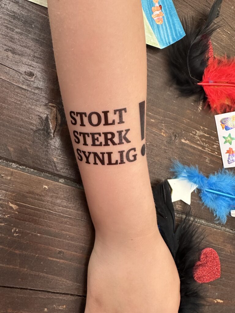 En barnearm med en midlertidig tatovering med teksten "Stolt, sterk, synlig!". Rundt armen ser vi papirhjerter, stjerner og fjær som brukes til å lage kunst. 