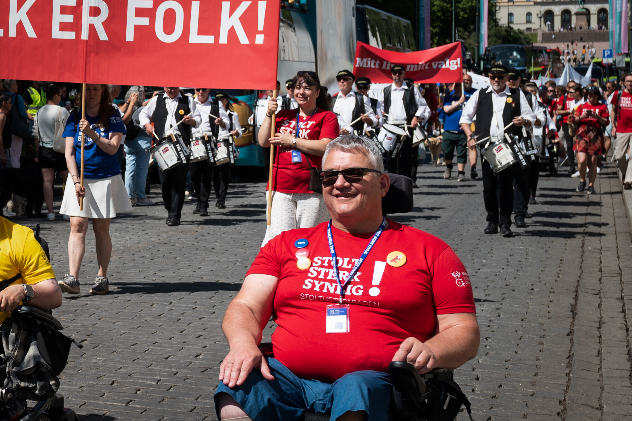Aage Gjesdal foran parolen "Folk er folk" i paraden i år. Han har rød t-skjorte med teksten "Stolt, sterk, synlig!" på. Han sitter i en rullestol.