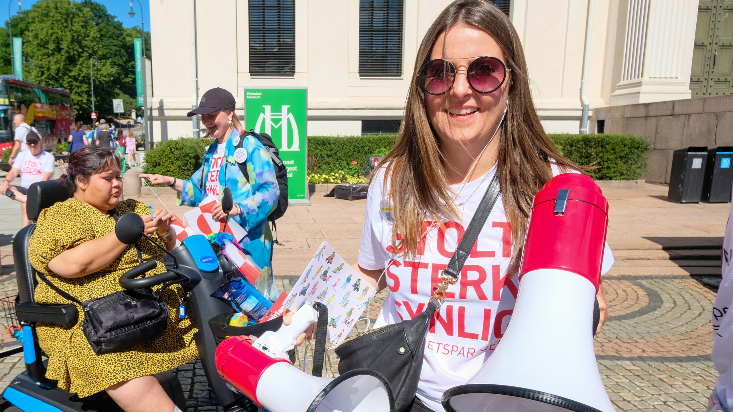 Anneth Nielsen med en stor ropert i hånden, et stort smil, solbriller og en hvit t-skjorte med teksten "Stolt, sterk, synlig!". Hun har brunblondt hår.