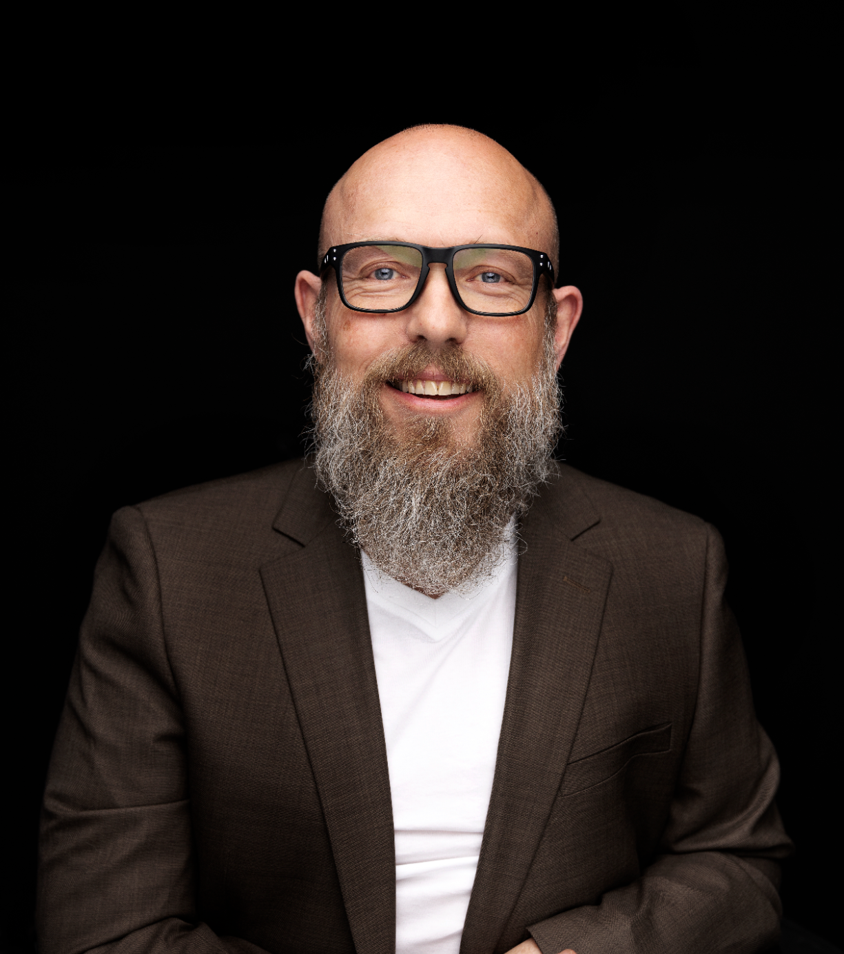 Stig Morten Skjæran, har på briller, langt grått skjegg. Har på en brun dressjakke og hvit t-skjorte under. 
