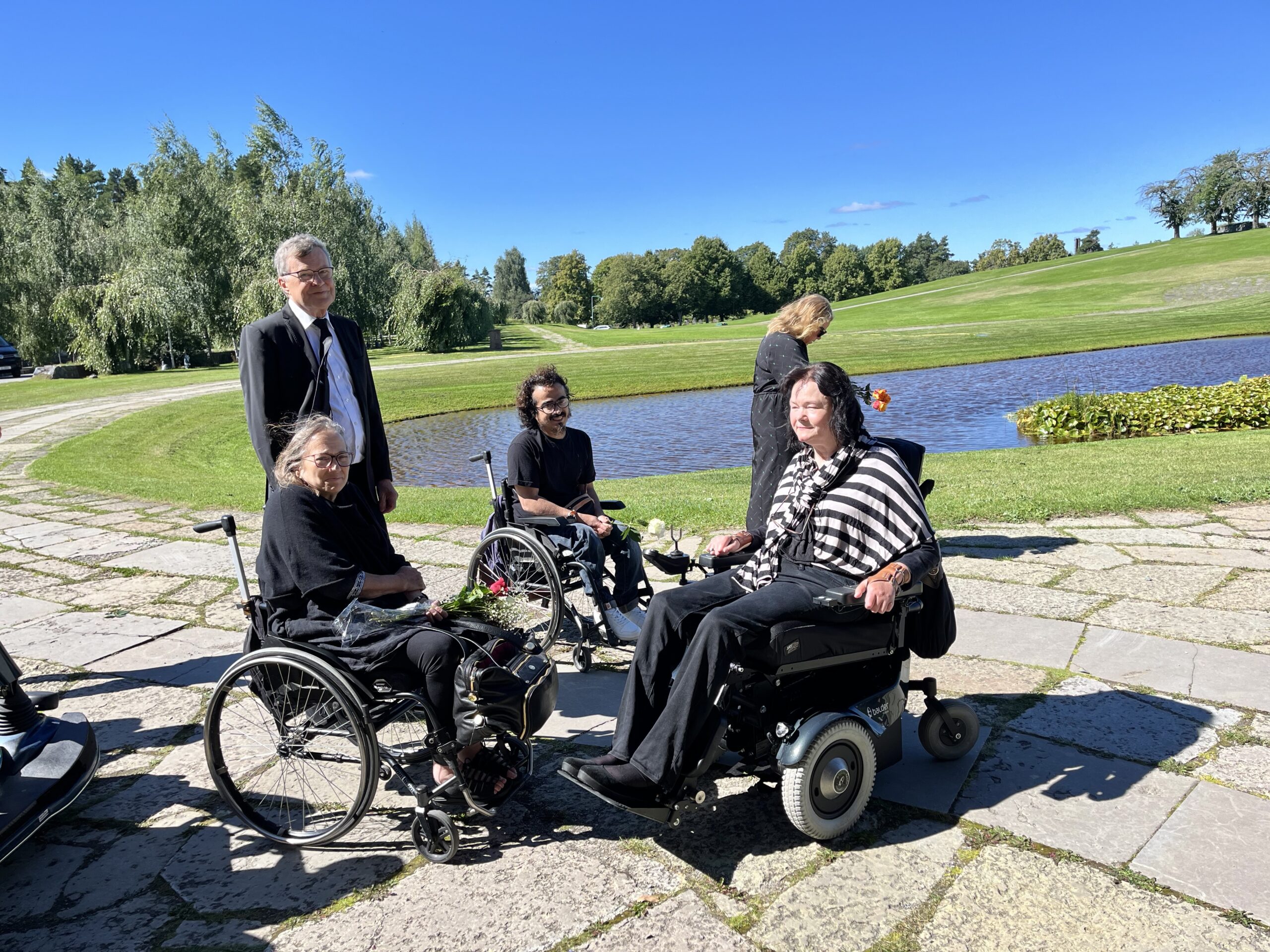 Biletet viser eit grønt område med dam i bakgrunnen, og i forgrunnen ei lita klynge menneske i rullestolar, ei av dei Vibeke Marøy Melstrøm, og nokre gåande. Samtlege er kledd i svart i høve gravferda.