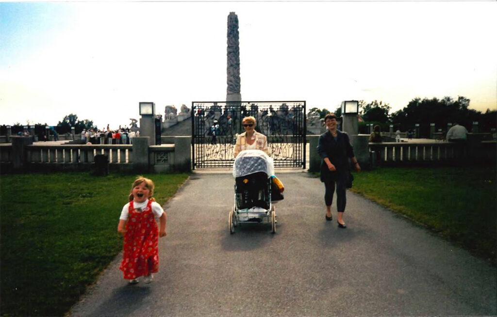 En liten veldig glad Mone med musefletter foran Monolitten i Frognerparken. Hun har på seg rød sommerkjole, hvit t-skjorte og smiler bredt tll kameraet. Hun er til venstre i bildet. Til høyre for henne ser vi en dame med barnevogn og en uten. Alle tre smiler til kameraet.