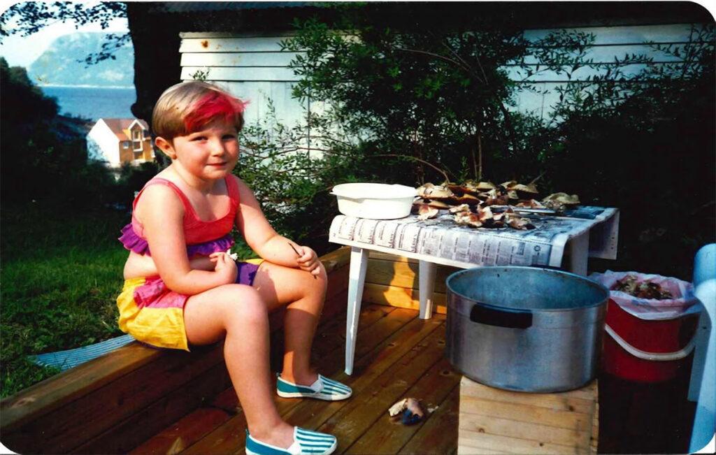 Mone som ung. Hun sitter på en terrassekant og smiler til kameraet. Hun har på seg en fargeglad short og matchende bikinioverdel med rysjer på. Hun har rød lugg og blå og hvite sko. På bordet ved siden av ligger det en haug med krabber, og ved siden av står det en stor gryte..Det er sol.