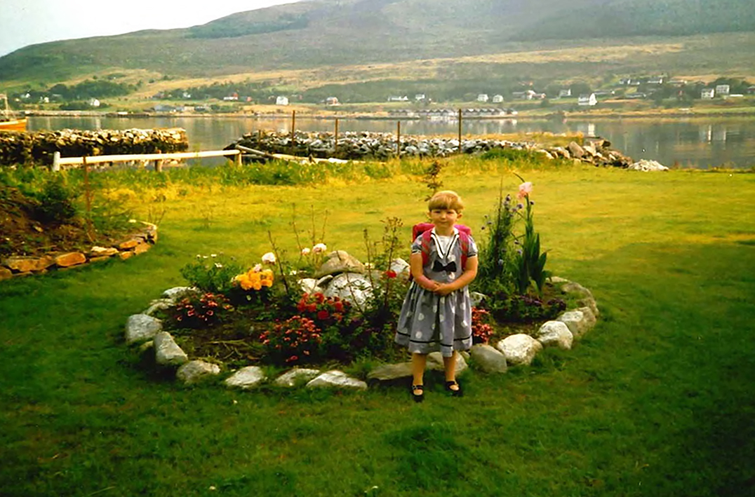 Mone er seks år. Hun står i en vakker hage med blomster og grønt. Bak henne ser vi fjell og fjord. Hun har klassisk bollesveis, grå kjole med hvite prikker, svarte ballerinasko og rosa skolesekk. Hun smiler til kameraet. Den høyre armen er mindre enn den venstre.