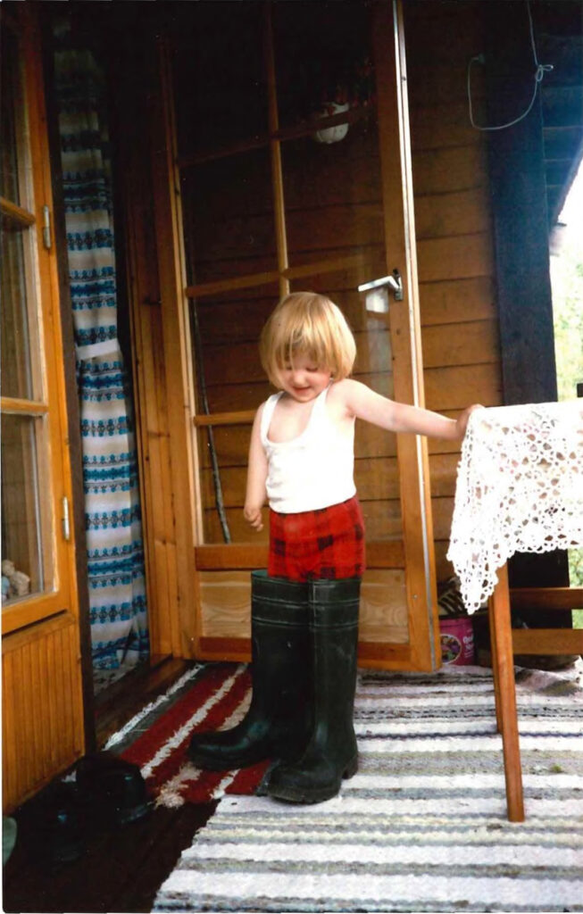 Mone som liten. Hun har på seg alt for store gummistøvler, en rød bukse og hvit topp. Hun har blond bolleklipp og står på fargerike filleryer ved siden av et bord med blondeduk. Hun smiler mens hun ser ned på støvlene.