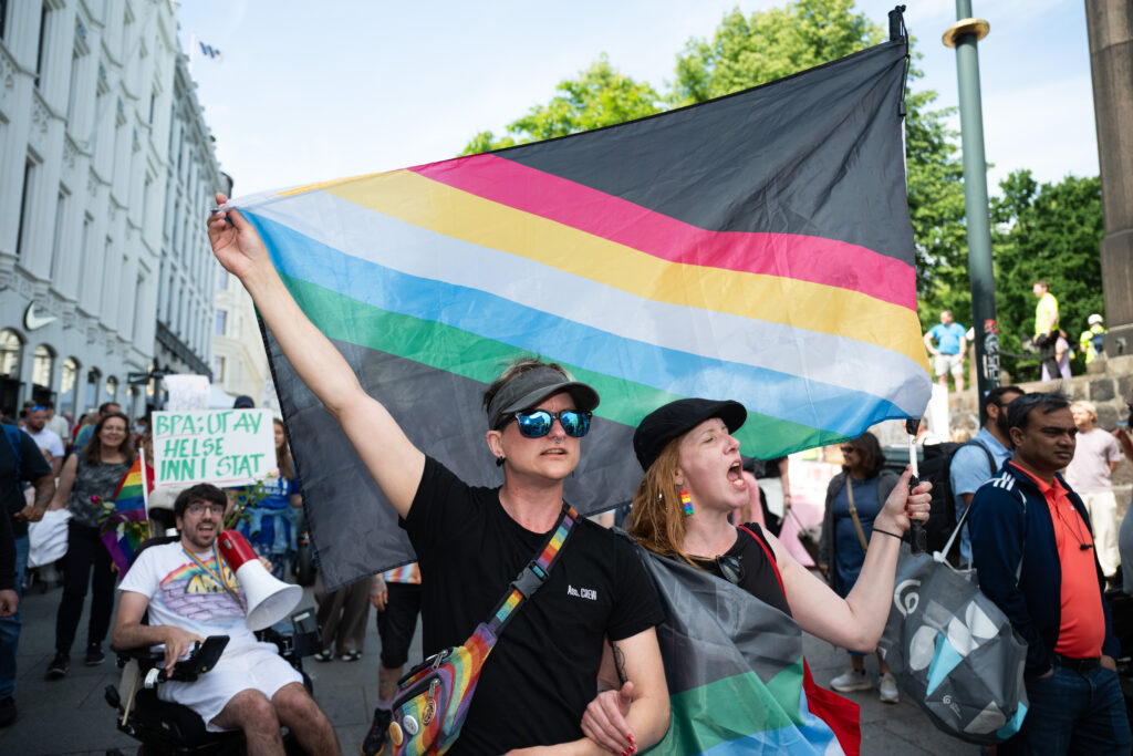 Bildet er tatt i Karl Johans gate i Oslo under Stolthetsparaden 2025. To mennesker holder opp et stort Disability Pride-flagg mens de roper slagord ut til folkemassen. Flagget har sort bakgrunn og fem striper i ulike farger, grønt, blått, hvitt, gult og rødt, diagonalt over bakgrunnen.