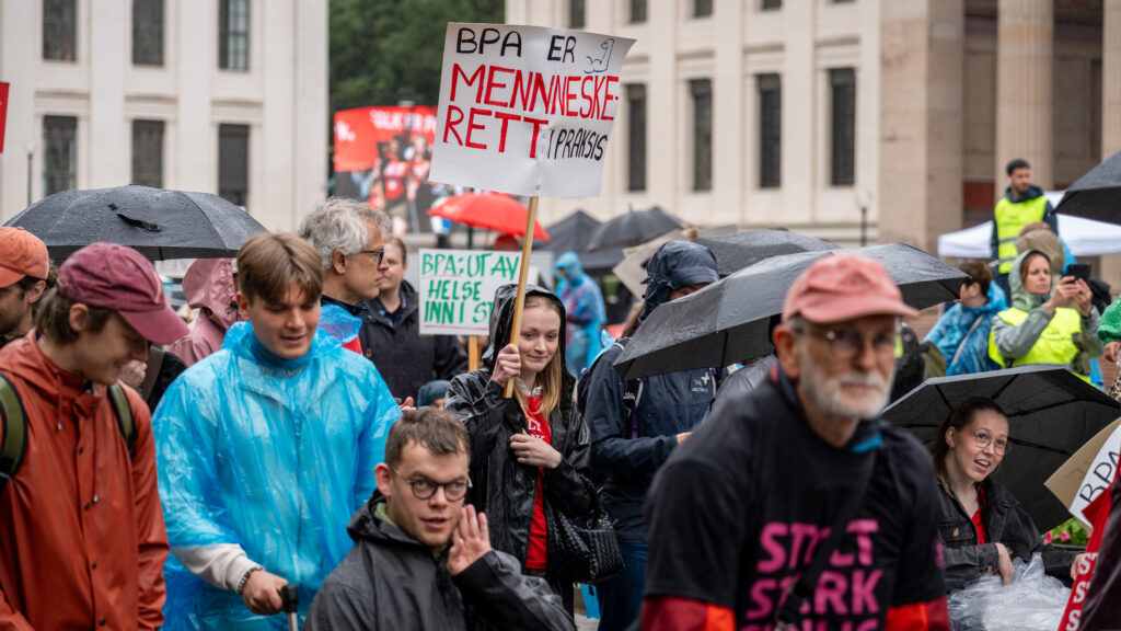 Bildet viser regnkledde paradegjengere i flokk nedover Karl Johan. Midt i bildet holder en blond ung kvinne en parole med ordene "BPA ER MENNESKERETT I PRAKSIS".