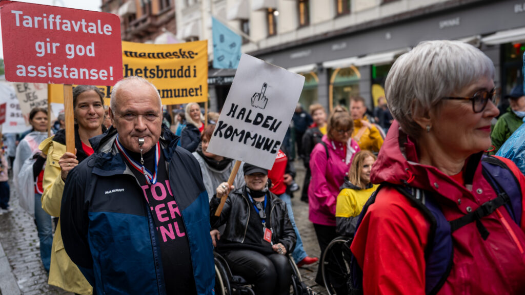 En eldre punker-kvinne holder en parole som viser fingeren til Halden kommune. Bak henne skimtes en gul parole med ordene "Stopp menneskerettighetsbrudd i norske kommuner".