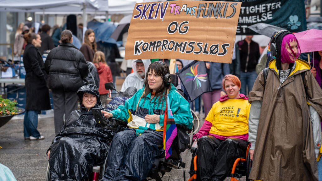 Aktivist og Uloba-ansatt Ihra Lill Scharning ikledd regntøy og med prideflagg og en stor papirbrun parole med teksten: SKEIV, TRANS, FUNKIS OG KOMPROMISSLØS.