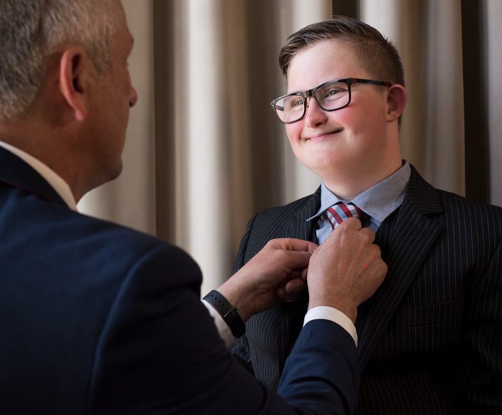 En person i mørkstripet dress, skjorte og rutete slips står mens en annen person justerer slipset. Begge er kledd formelt. Bildet er tatt innendørs foran lyse gardiner.